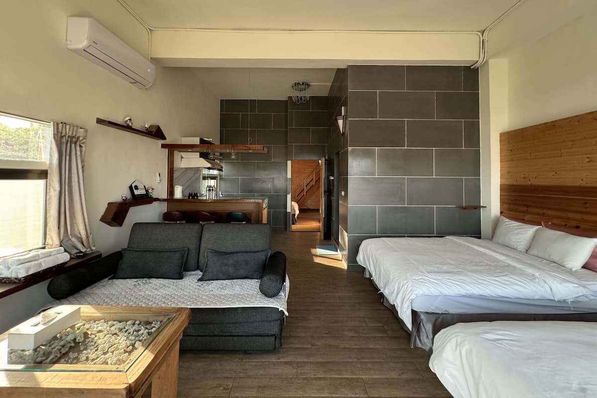 A modern bedroom with two beds, a sofa, a small table with a decorative item, and a view into the kitchen area.