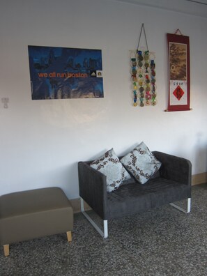 Una habitación con un sofá gris, dos almohadas estampadas, un pequeño taburete beige, una pared con un cartel, un tapiz de medallas y un cuadro enmarcado.