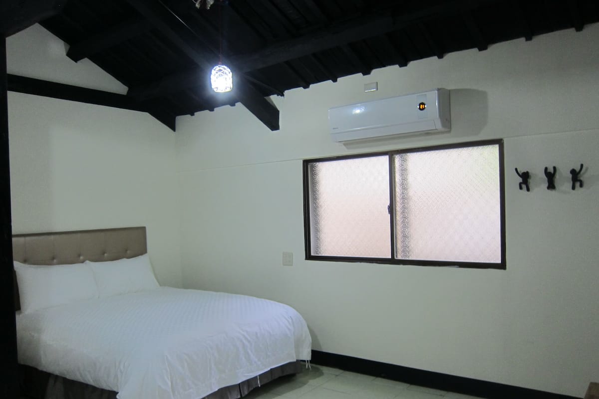 A bedroom with a bed, a window with frosted glass, an air conditioner, and a wall-mounted shelf with three figurines.