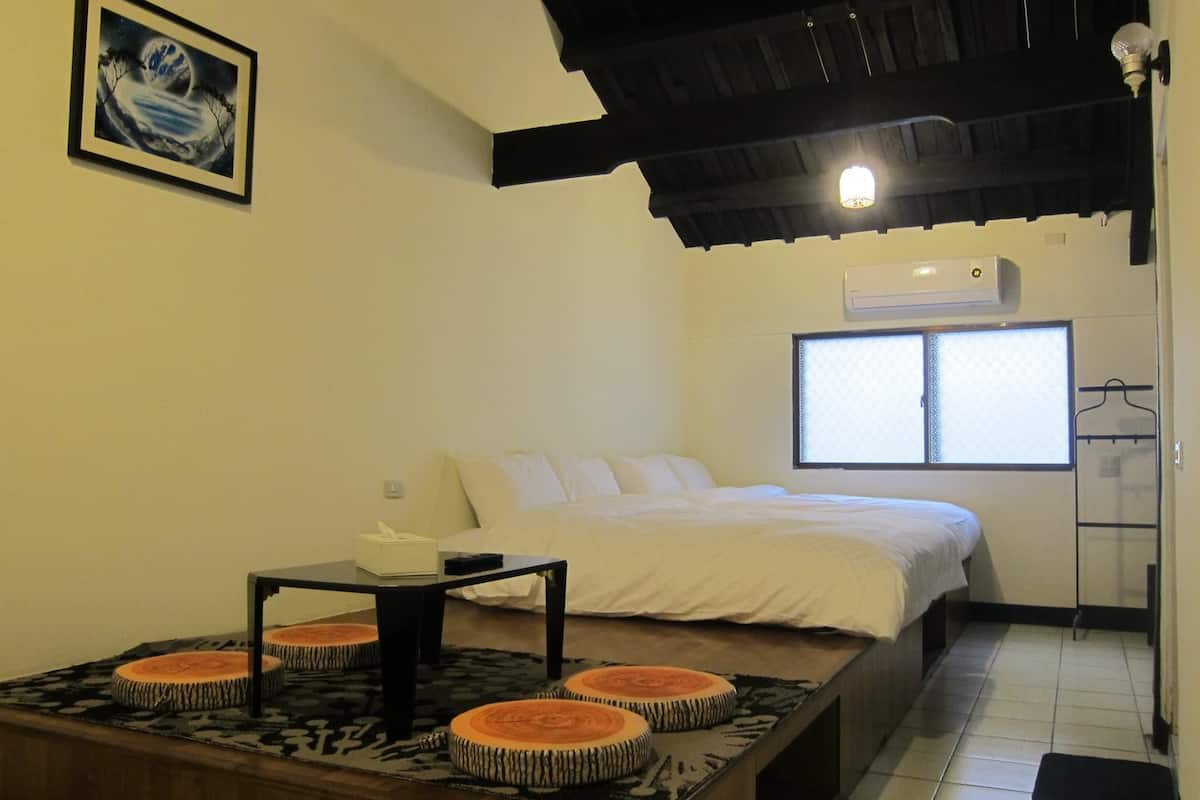 A hotel room with a bed, a small table with a tissue box, a window with blinds, and a wall-mounted air conditioner.