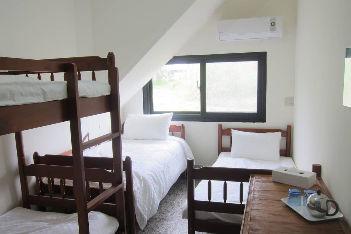 A dormitory room with bunk beds, a single bed, a wooden desk with a teapot and cups, and a window with a view of greenery.