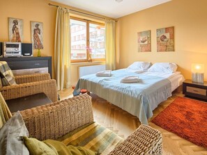 Une chambre confortable avec un lit, deux fauteuils en osier, une petite table, un radiateur et des œuvres d’art accrochées aux murs.