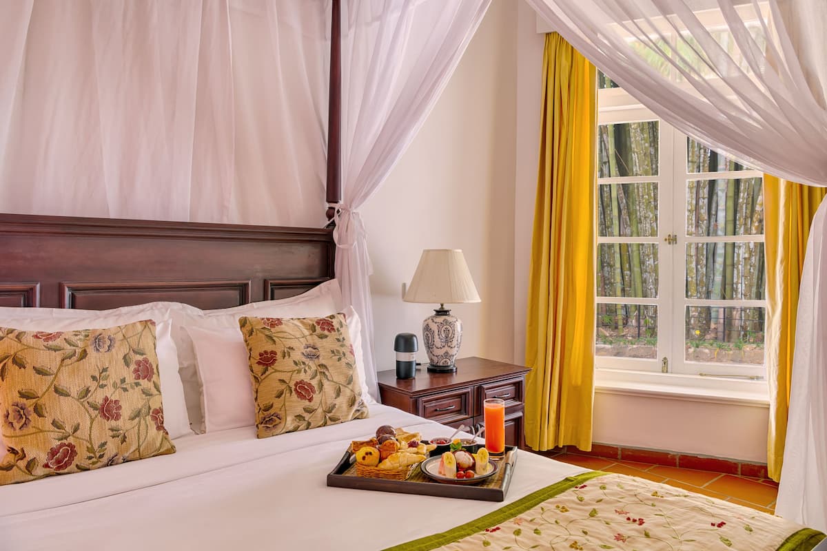 Una cama bien tendida con almohadas florales, una bandeja de desayuno, una lámpara de noche y una ventana con cortinas amarillas.