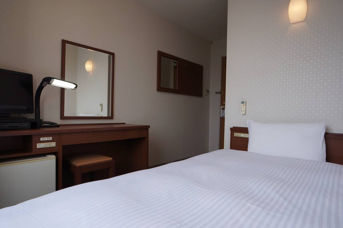 Habitación de hotel con cama, escritorio con lámpara, espejo, teléfono y mesita de noche con almohada.
