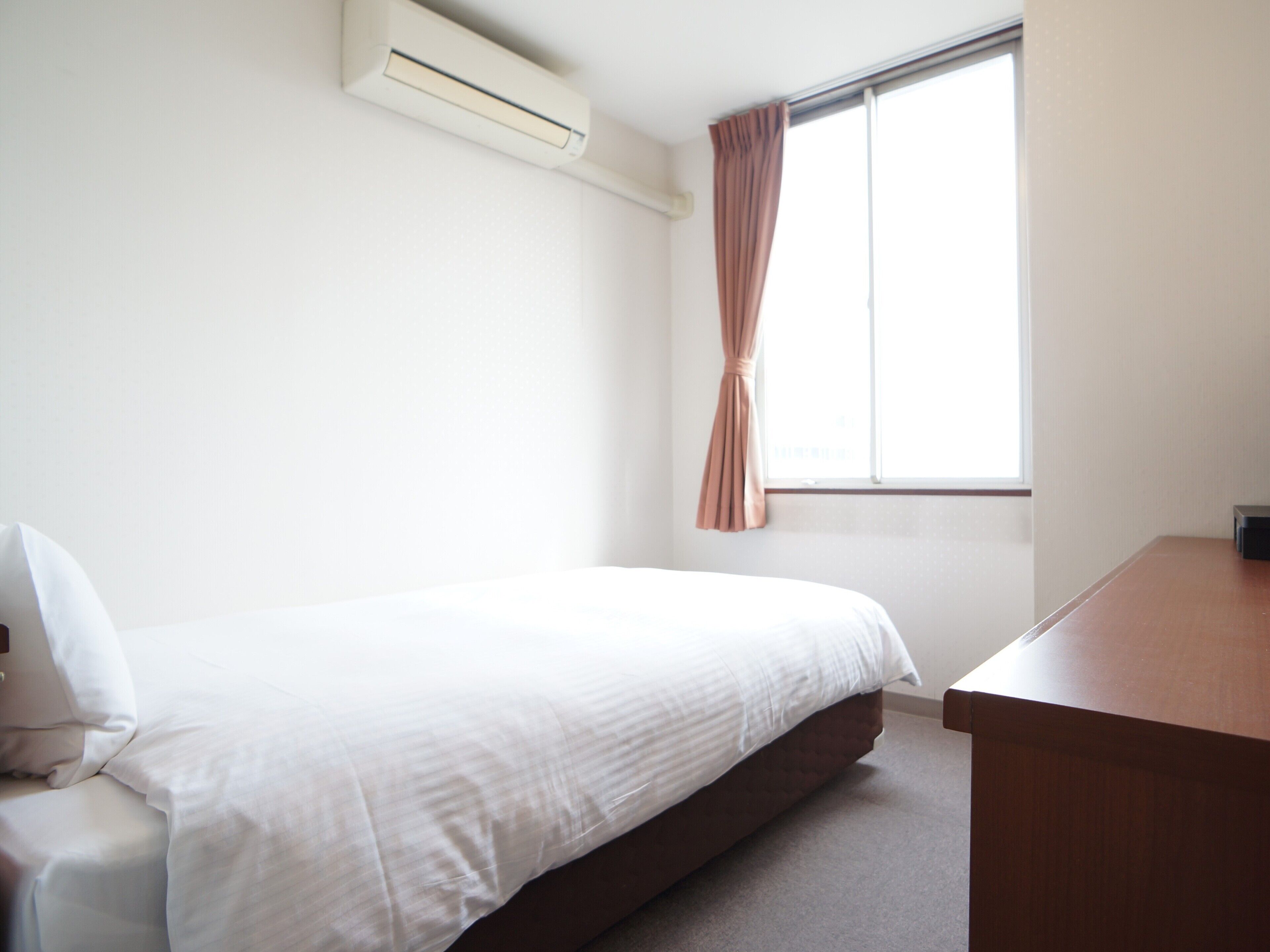 Ein schlicht eingerichtetes Hotelzimmer mit einem Einzelbett, einem Holztisch und einem Fenster mit Vorhängen.