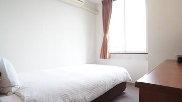 Ein schlicht eingerichtetes Hotelzimmer mit einem Einzelbett, einem Holztisch und einem Fenster mit Vorhängen.