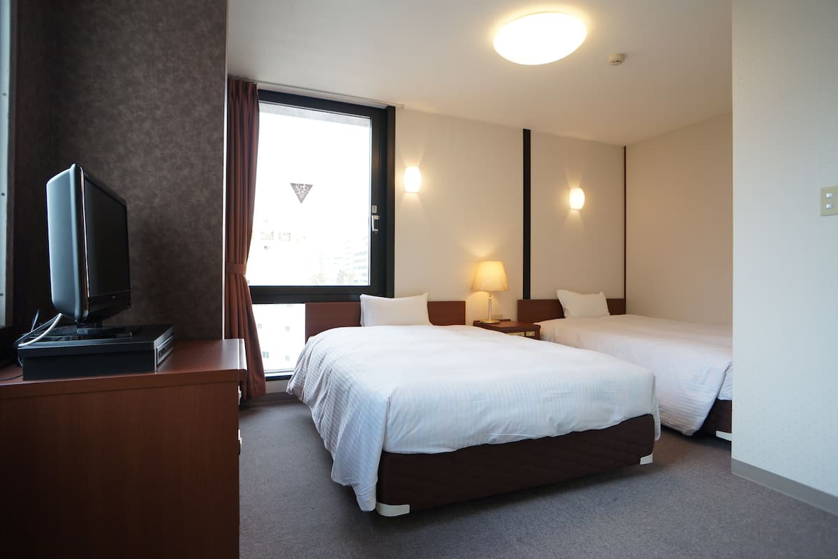 Una habitación de hotel con dos camas, un escritorio con televisor y una ventana con cortinas.