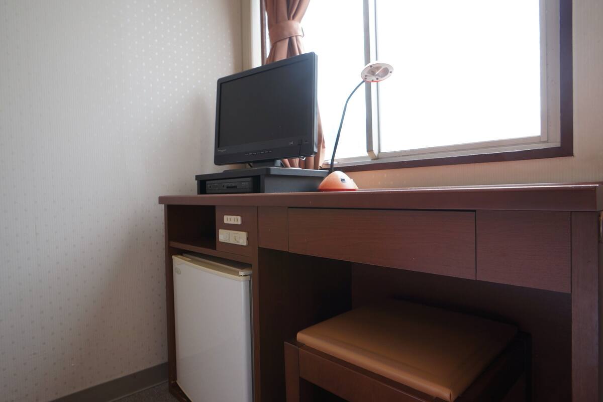 Una habitación compacta con un escritorio de madera, un refrigerador pequeño y una ventana con cortinas.