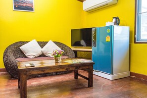 Ein Zimmer mit gelber Wand, einer Couch mit Kissen, einem Holztisch mit Fernbedienung und Zeitschrift, einem blauen Kühlschrank und einem Fernseher.