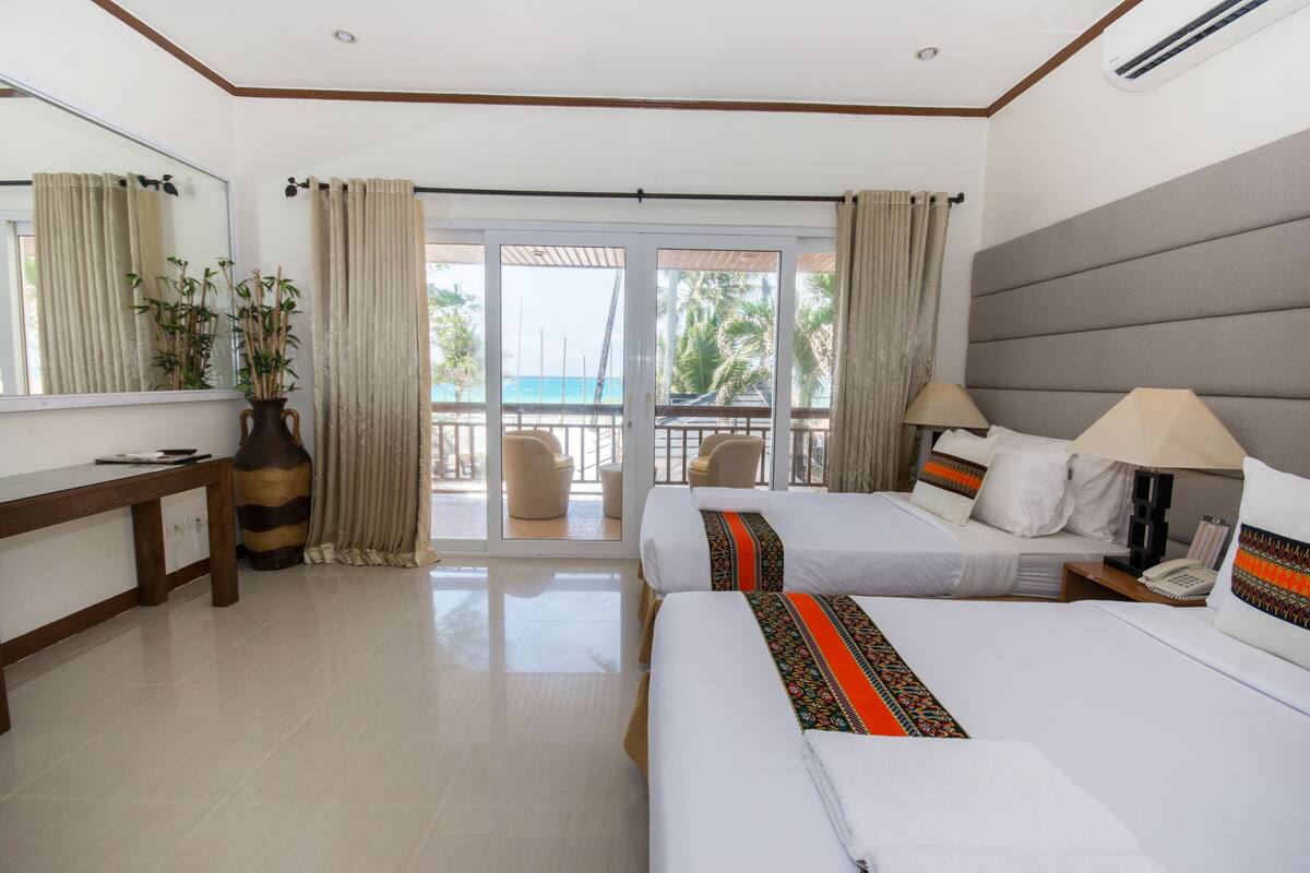 Une chambre d’hôtel avec deux lits, un grand miroir, un bureau, un vase et une vue sur la plage, visible à travers les portes vitrées.
