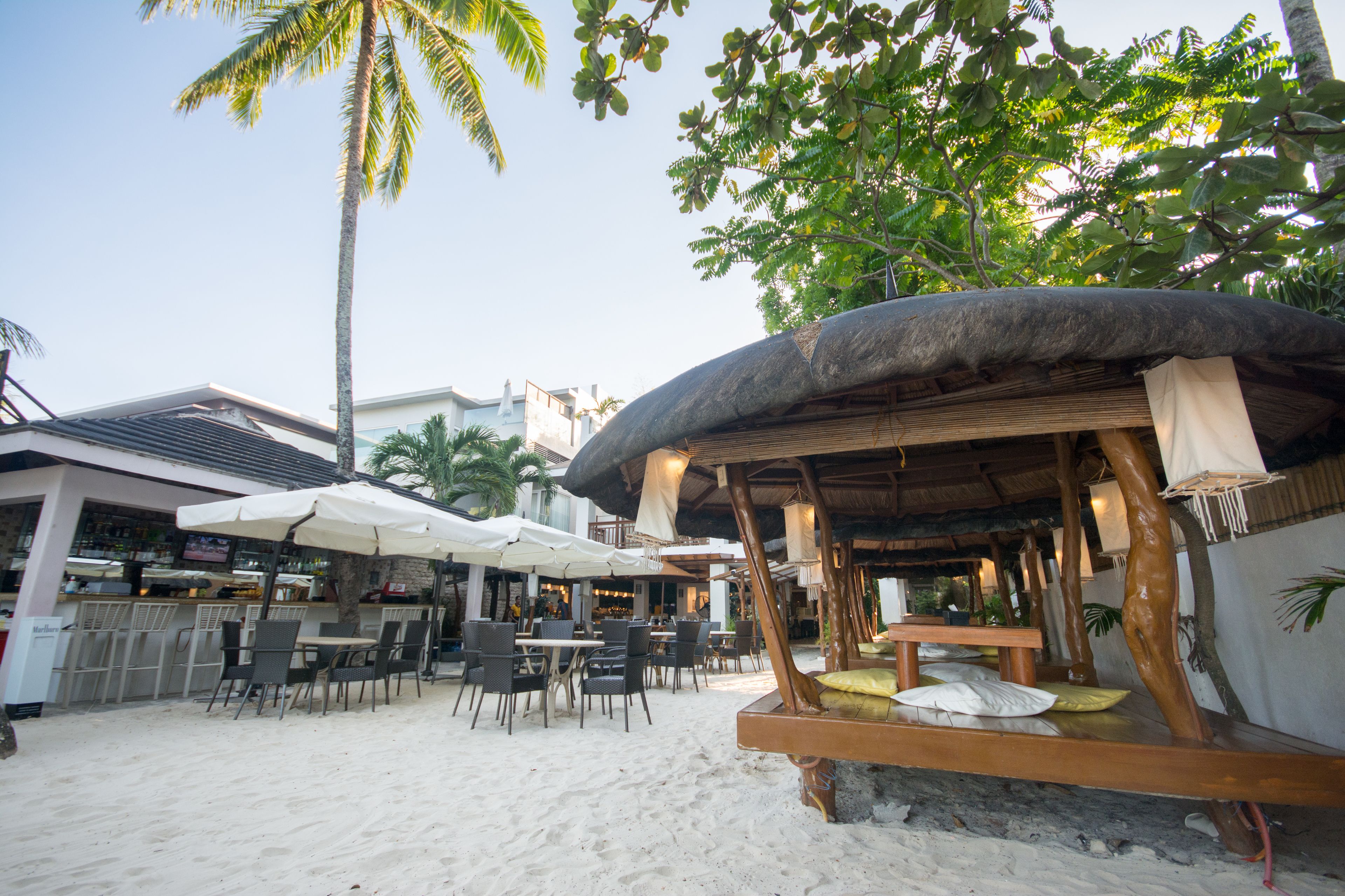 A beachside restaurant with outdoor seating, palm trees, and a thatched-roof hut.