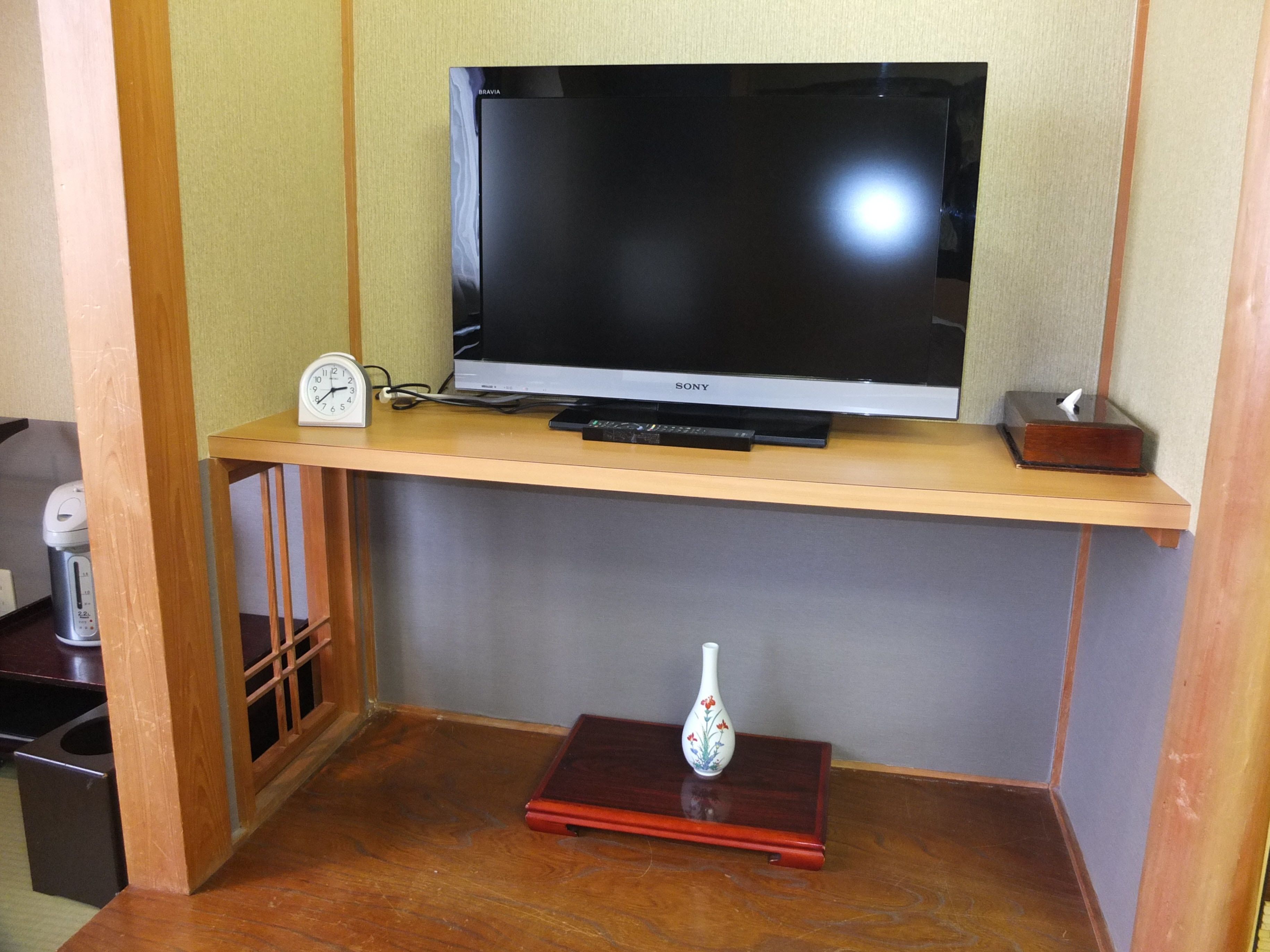 A flat-screen TV on a wooden shelf, a clock, a remote control, and a decorative vase on a small table.
