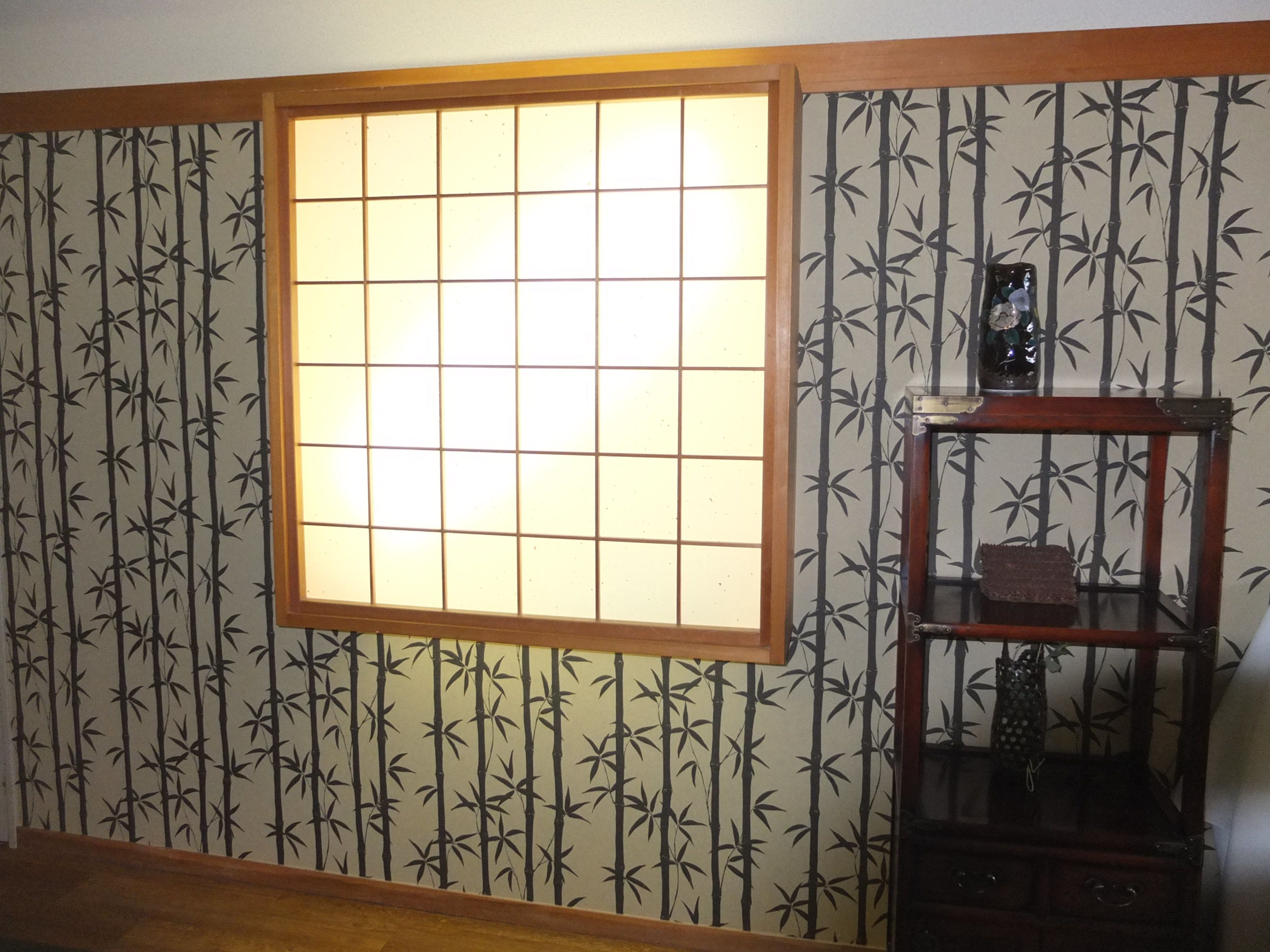 A room with a shoji screen window, bamboo wallpaper, and a traditional wooden cabinet with a glass top.