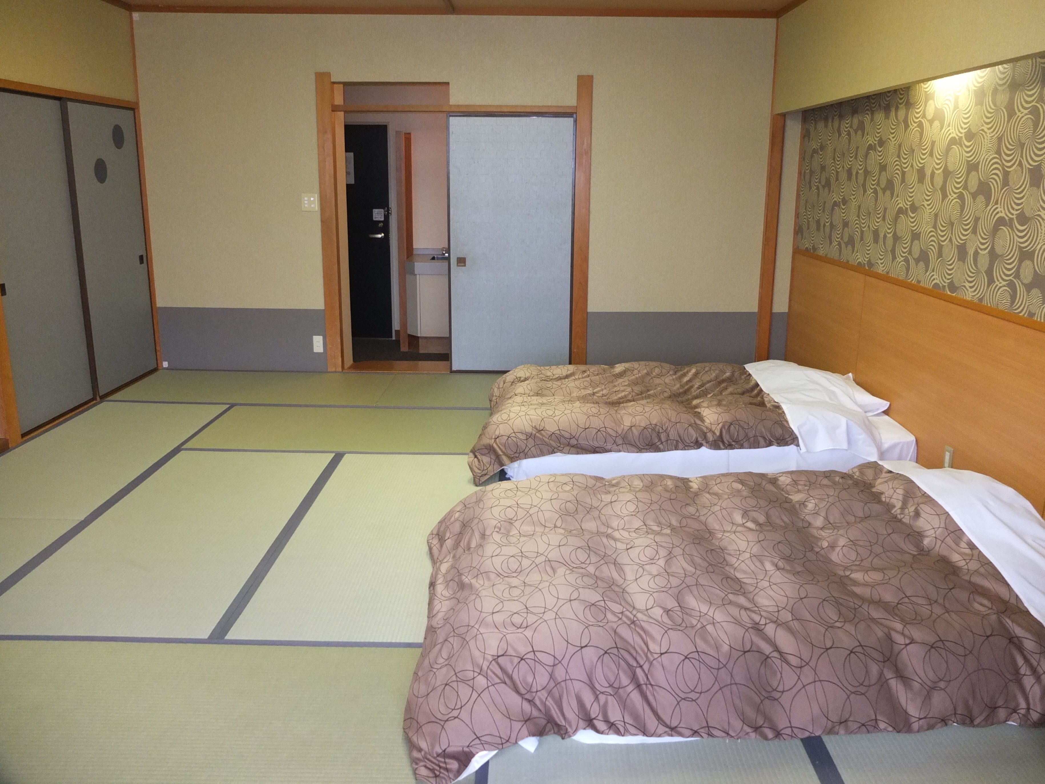 Ein Zimmer mit Tatami-Bodenbelag, zwei Betten mit braunen, gemusterten Bettwäsche, einem Holz-Kopfteil und einer Tür zu einem Badezimmer.