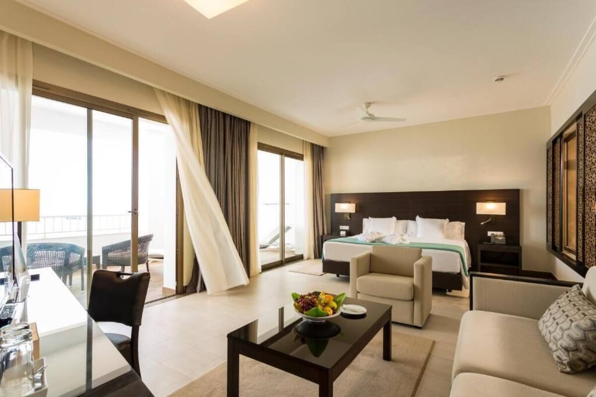 A hotel room with a bed, a sofa, a dining table, and a balcony with outdoor seating.