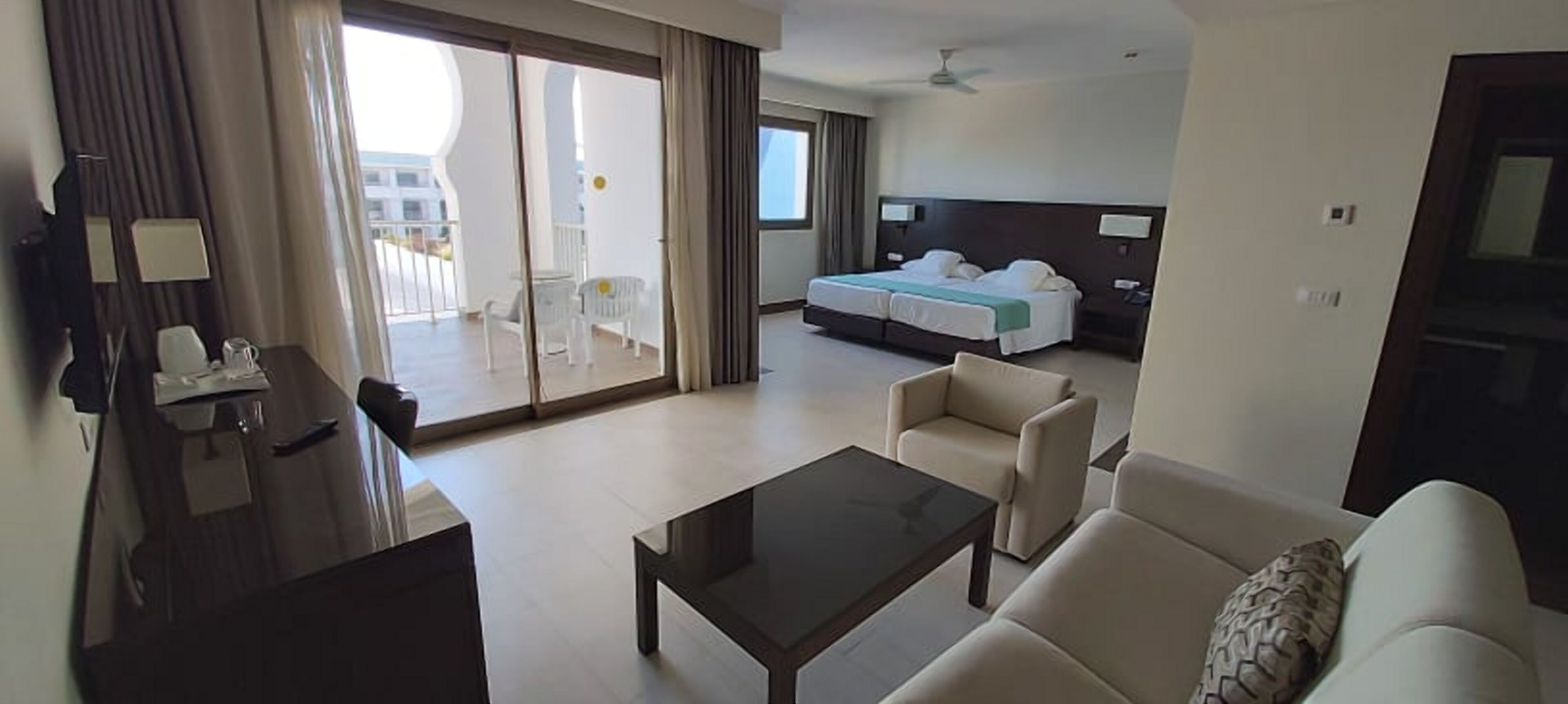 A hotel room with a balcony, a bed, a sofa, a coffee table, and a desk with a chair.