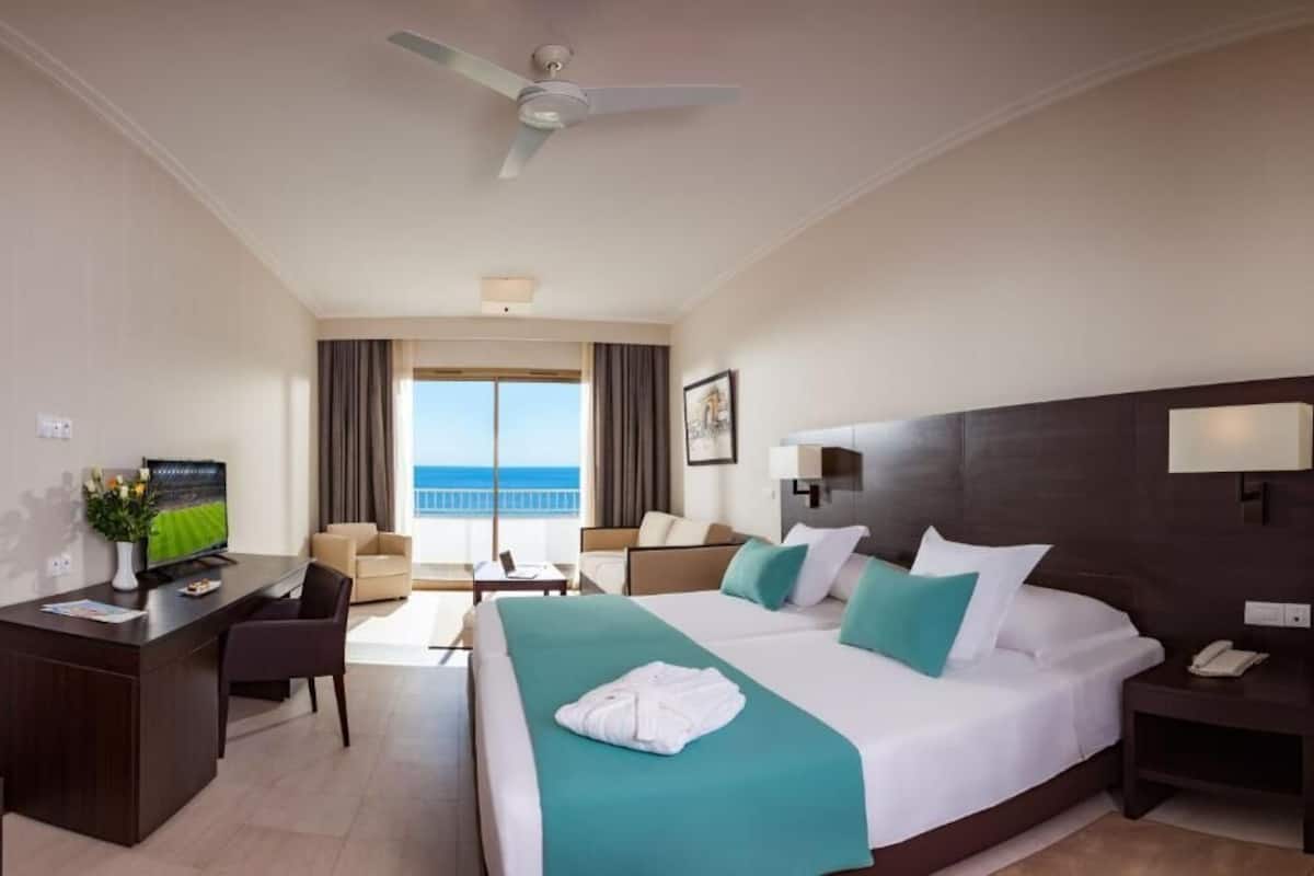 A hotel room with a bed, desk, chair, and a view of the sea.