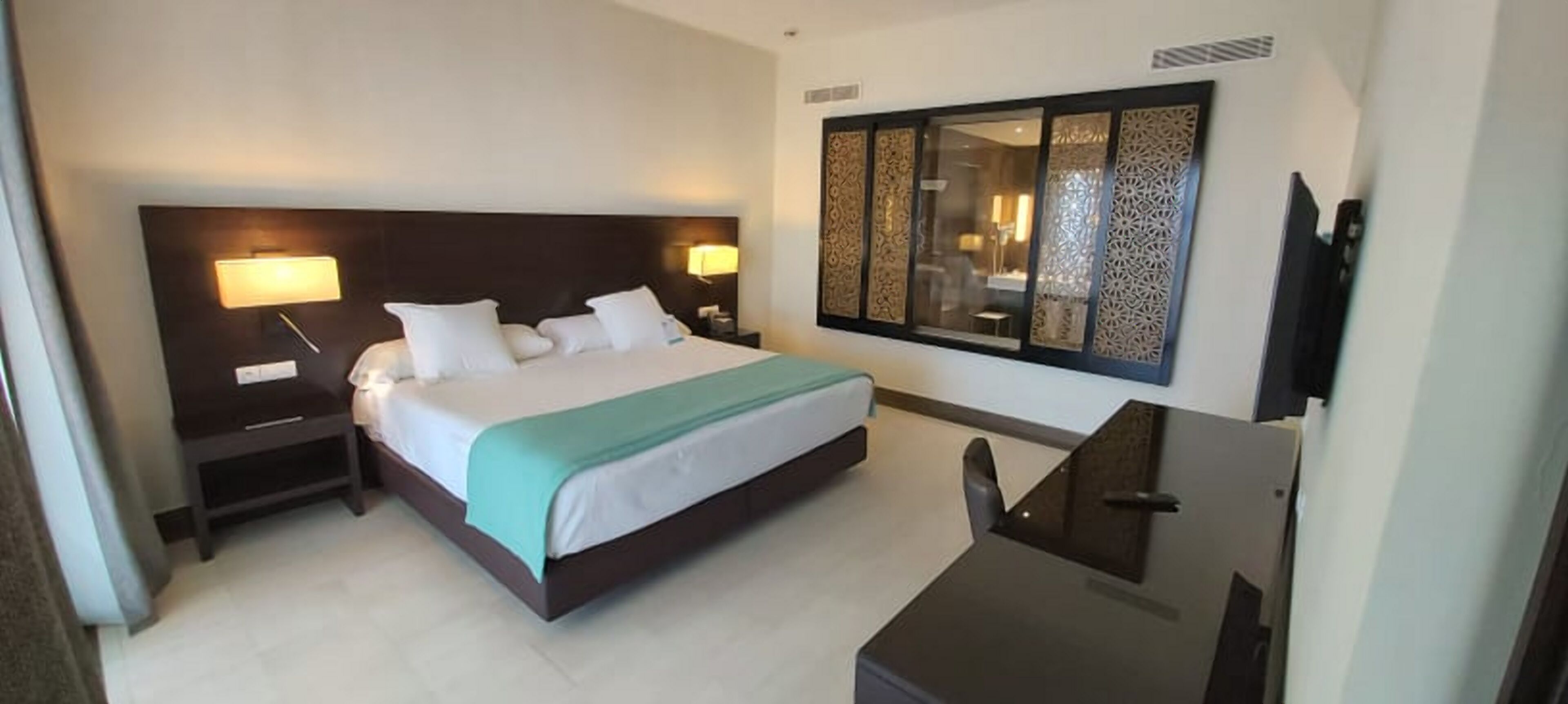 Une chambre d’hôtel avec un grand lit, deux lampes de chevet, un bureau et une grande fenêtre ornée de panneaux décoratifs.