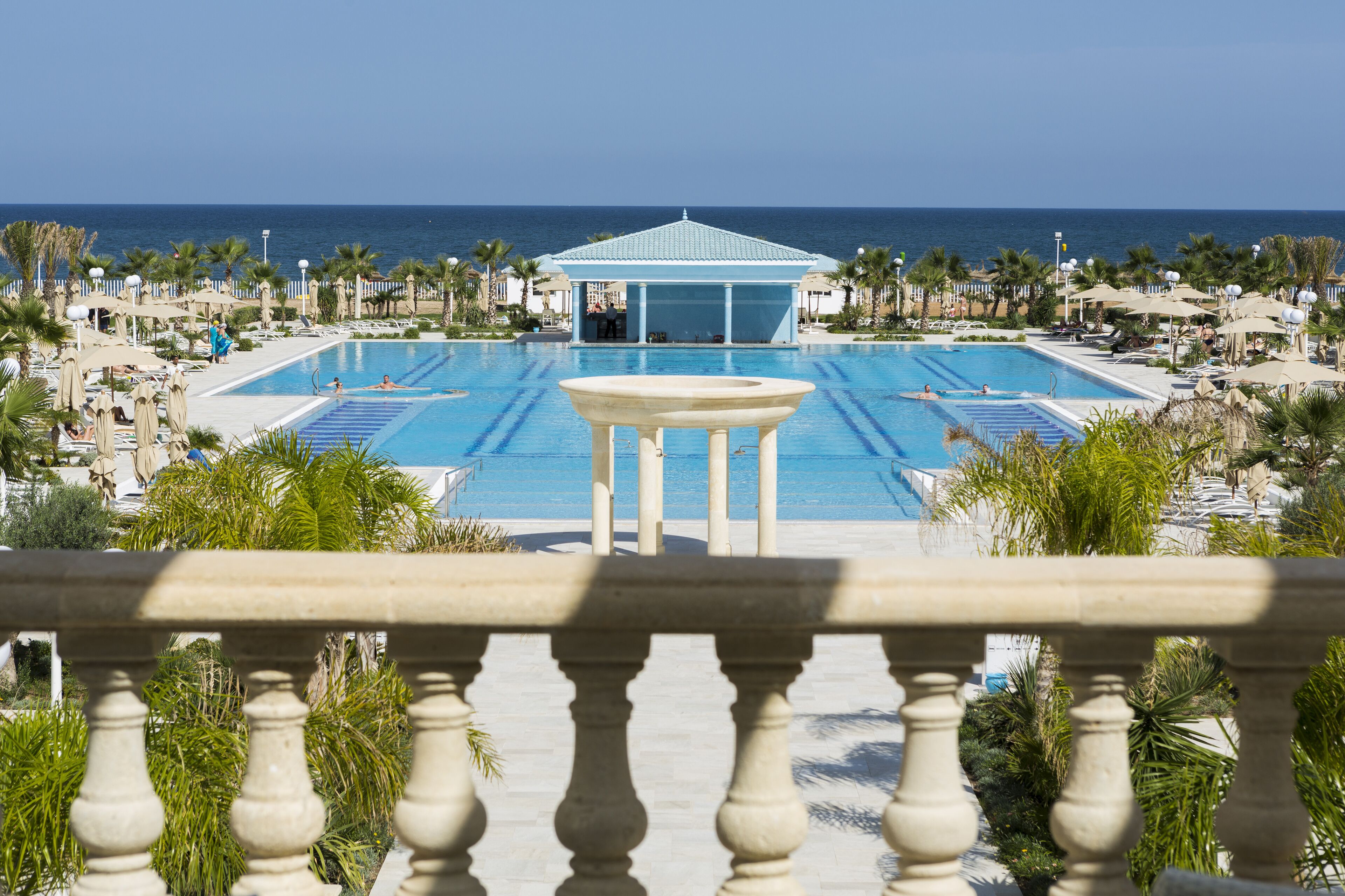 A large outdoor swimming pool with a gazebo in the center, surrounded by palm trees and lounge chairs.