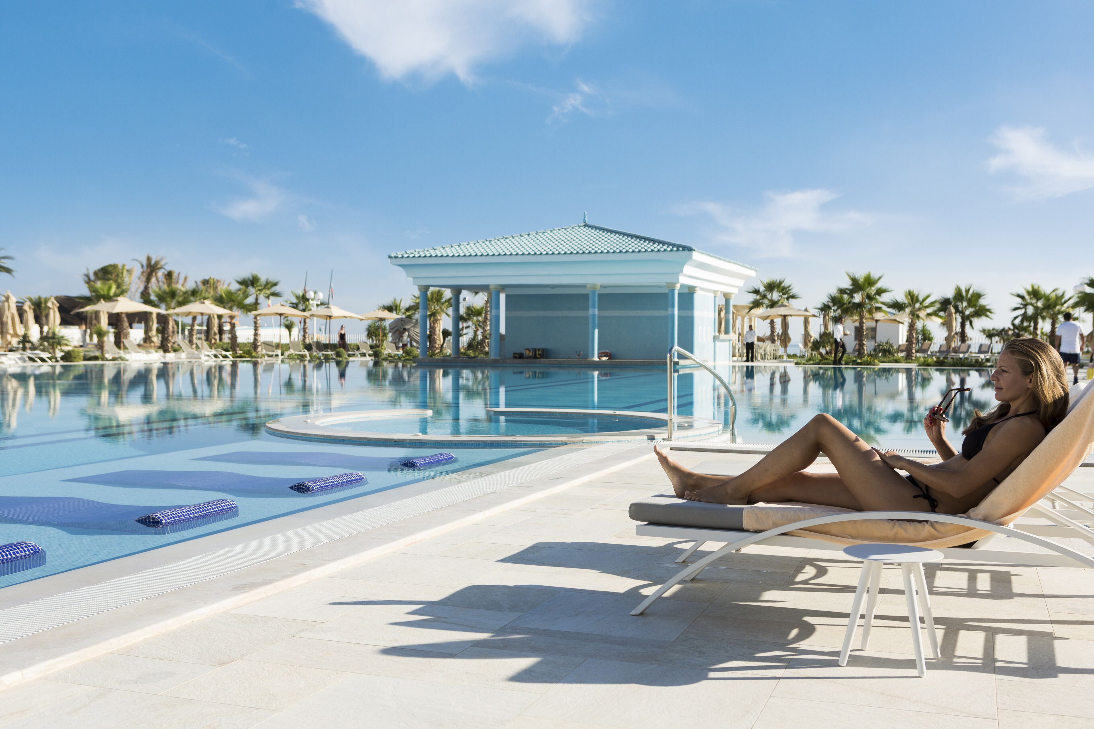 Un espace aménagé au bord de la piscine, avec des chaises longues, un coin salon abrité et des palmiers.