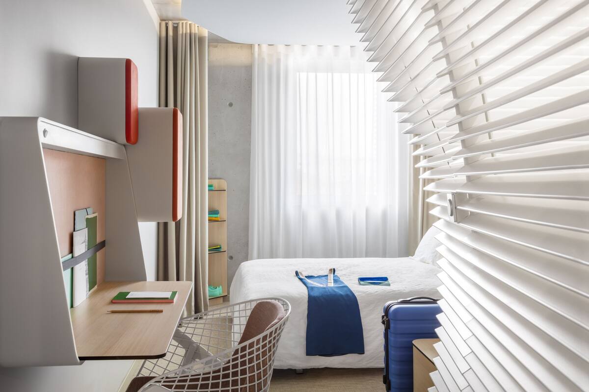 A hotel room with a bed, a desk, a chair, and a window with blinds.
