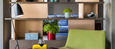 A modern living room with a green chair, a small table with a glass of wine, and a bookshelf with various decorative items.