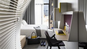 A modern hotel room with a bed, a desk, a chair, and a view of the city through large windows.