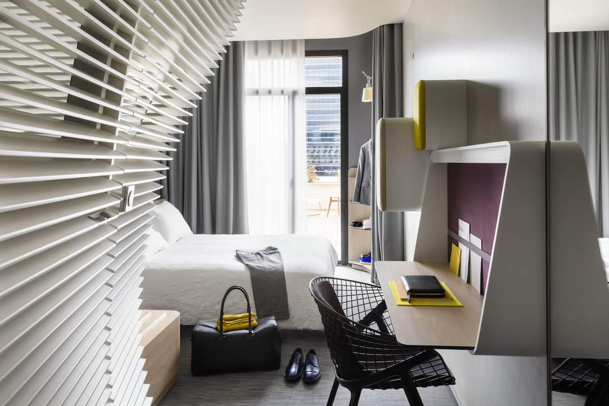 A modern hotel room with a bed, a desk, a chair, and a view of the city through large windows.