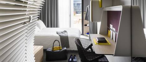 A modern hotel room with a bed, a desk, a chair, and a view of the city through large windows.