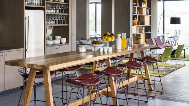 A modern kitchen with a long wooden table, bar stools, and hanging pendant lights.