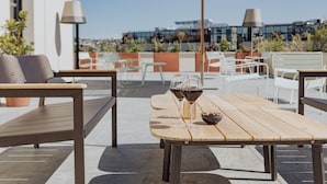 An outdoor seating area with a wooden table, two wine glasses, and a bowl of fruit. There are white chairs and a large umbrella.
