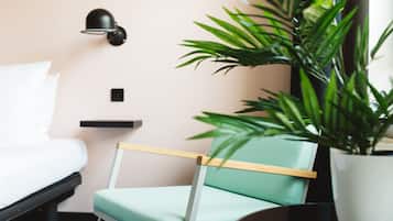 A room with a bed, a chair, a wall-mounted lamp, a small shelf, and a potted plant.
