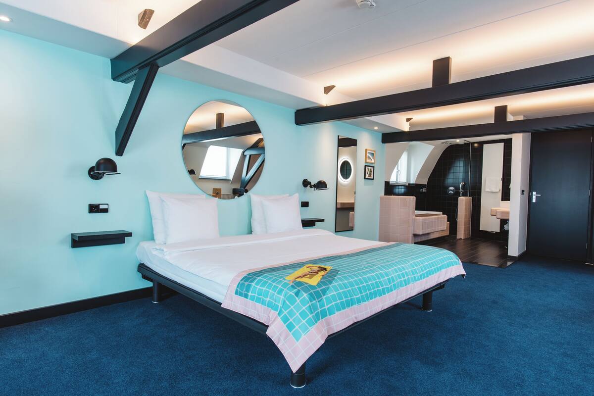 Ein modernes Hotelzimmer mit Bett, einem runden Spiegel, einer kleinen Sitzecke und einem durchgegangene Tür erreichbaren Badezimmer.
