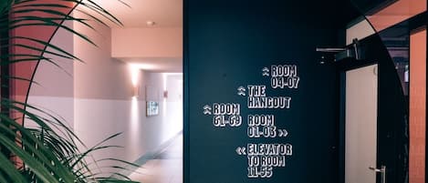 Un pasillo con indicaciones hacia varias habitaciones y un ascensor, una planta en maceta y un diseño interior moderno con una pared de acento roja.