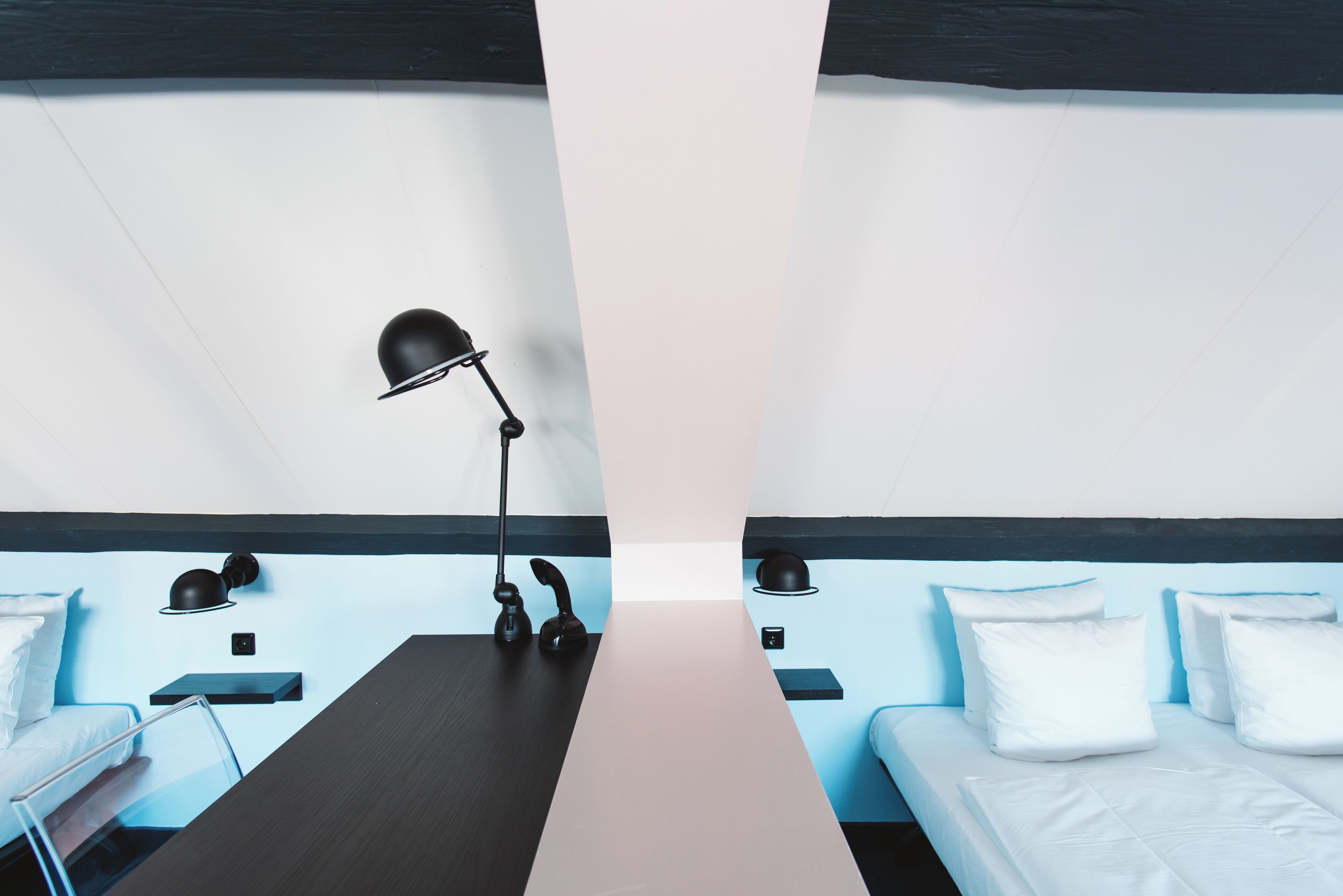 Una habitación de hotel moderna con un escritorio central, dos camas, una lámpara negra y una pared de acento azul.