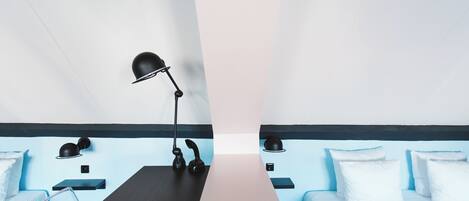 Una habitación de hotel moderna con un escritorio central, dos camas, una lámpara negra y una pared de acento azul.