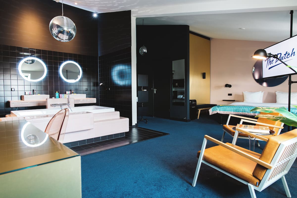Ein modernes Hotelzimmer mit einem großen Spiegel, runden Spiegeln, einer Discokugel, einem Bett mit grün-weiß kariertem Überwurf und einem Schreibtisch mit Lampe.