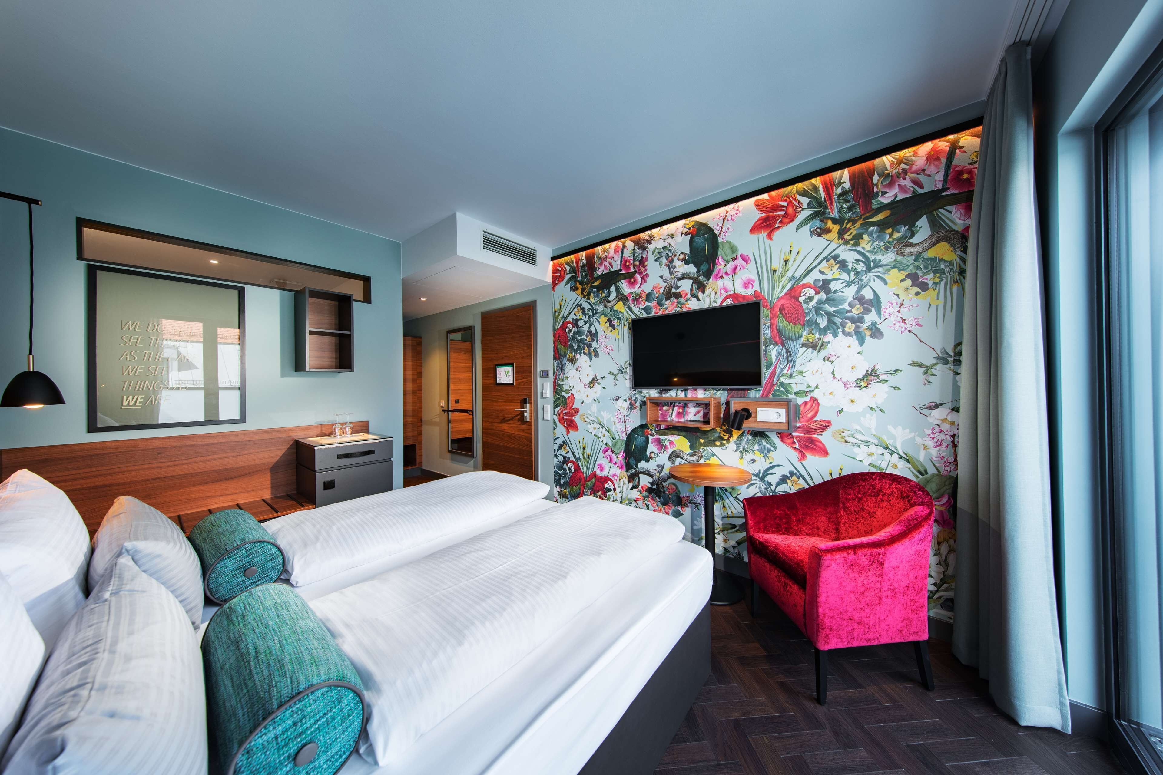 A hotel room with a large bed, a red armchair, a flat-screen TV, and a floral wallpaper.