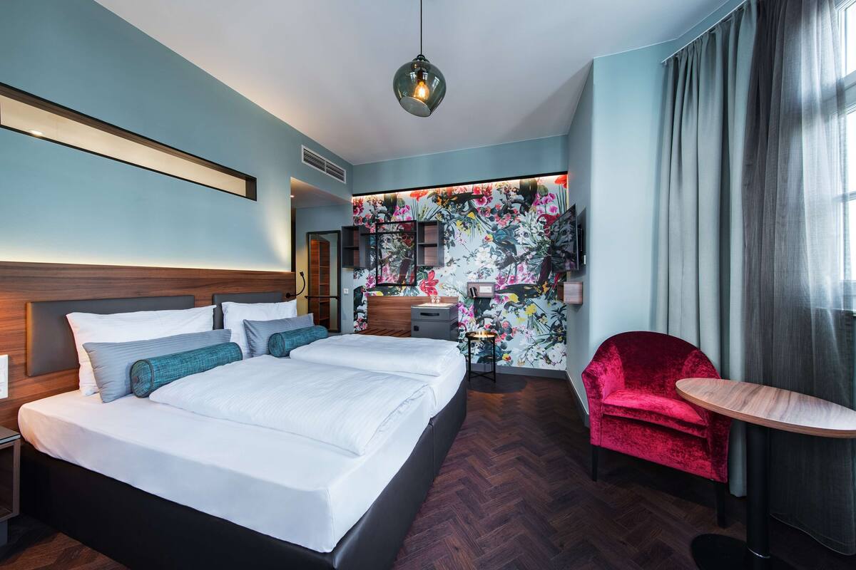 Una habitación de hotel moderna con una cama grande, una silla roja, una mesita pequeña y una pared decorada con estampado floral.