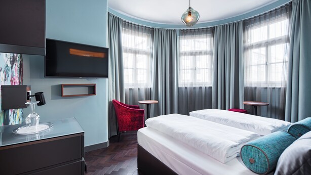 A modern hotel room with a large bed, a desk with a glass top, a red chair, and a TV mounted on the wall. - Maison Schiller (Munich)
