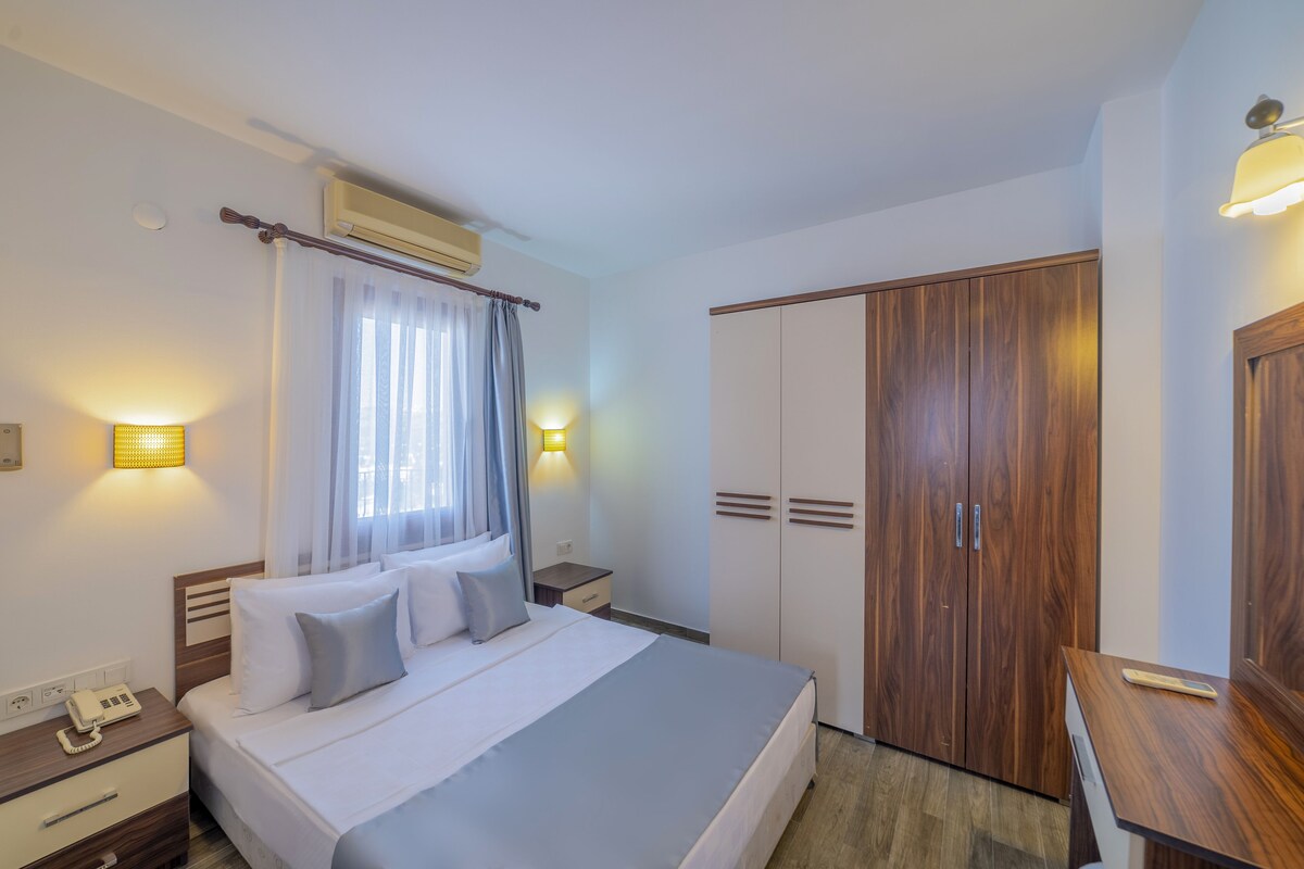 A hotel room with a bed, bedside table, lamp, wardrobe, and an air conditioning unit.
