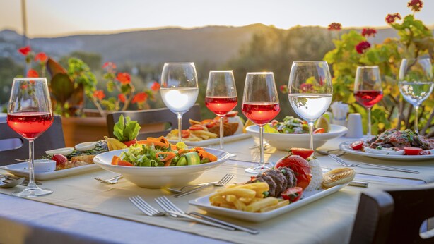 Una mesa dispuesta para una comida con diversos platos y copas de vino tinto, con vistas a un paisaje pintoresco.