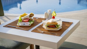 Una mesa de comedor con comida que incluye una hamburguesa y papas fritas, una ensalada y salsas, con vista a una piscina.