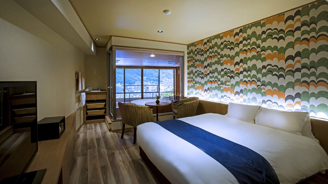 Ein Hotelzimmer mit einem großen Bett, einem Esstisch, zwei Stühlen, einer kleinen Küchenzeile und Blick durch ein großes Fenster.