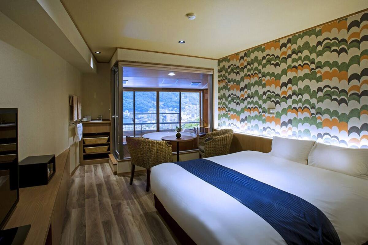 A hotel room with a large bed, a dining table, two chairs, a small kitchen area, and a view through a large window.