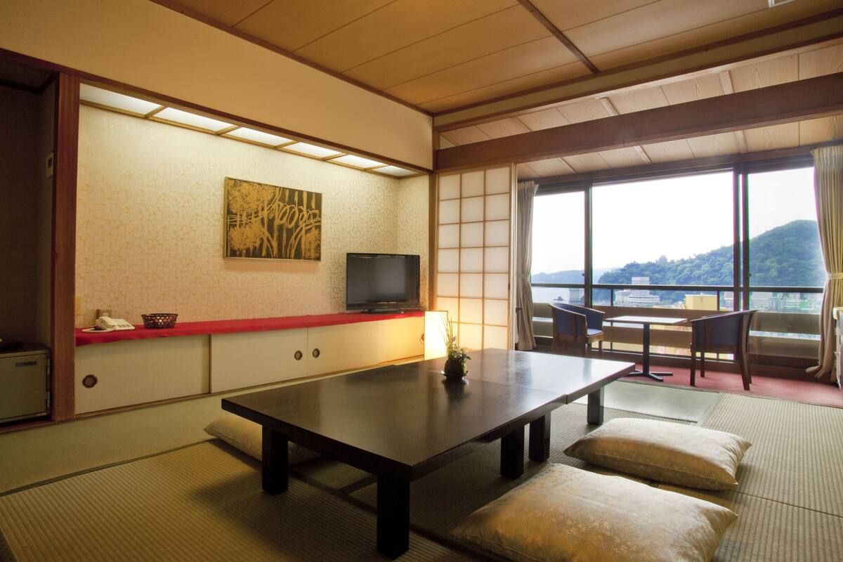 A traditional Japanese room with tatami flooring, a low table, cushions, a television, and a view of a mountain.
