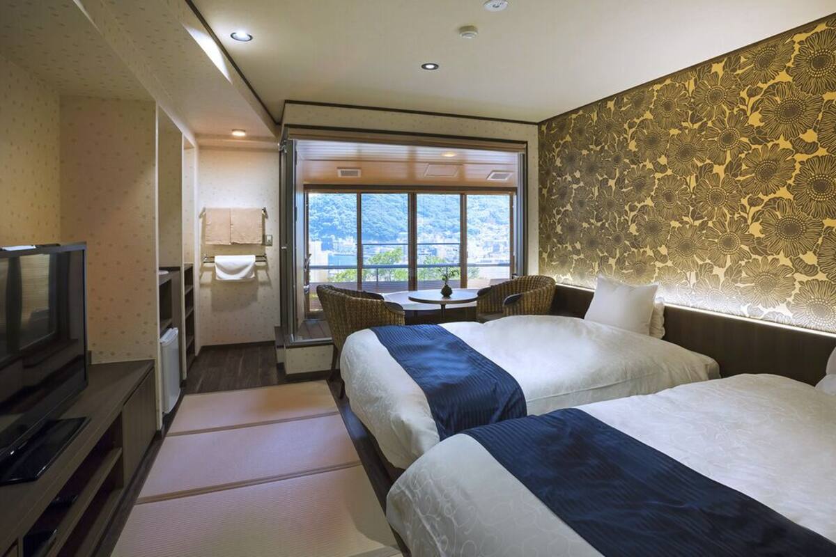 A hotel room with two beds, a TV, a small table, and a view of the outside.