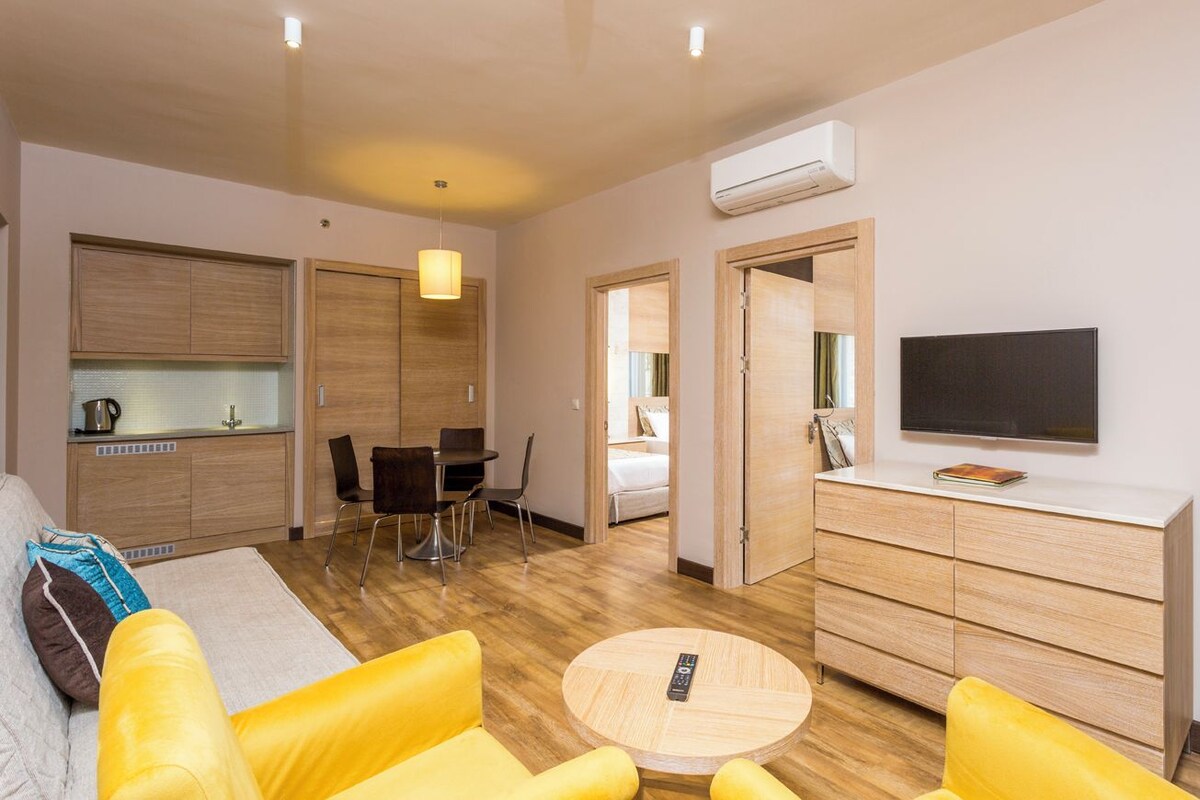 A modern hotel room with a sofa, a round coffee table, a TV, a wardrobe, and a kitchenette.