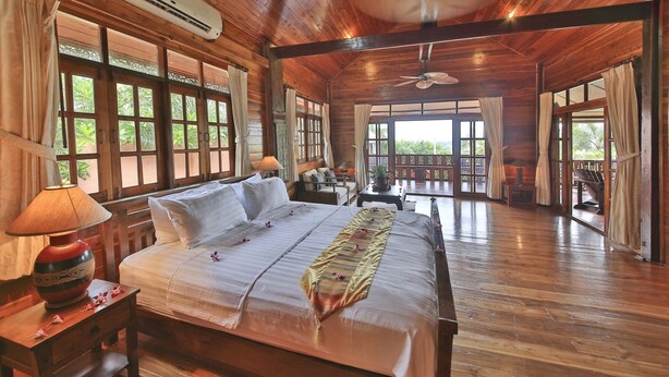 A spacious room with a large bed, wooden flooring, a ceiling fan, and large windows offering a view of the outdoors.
