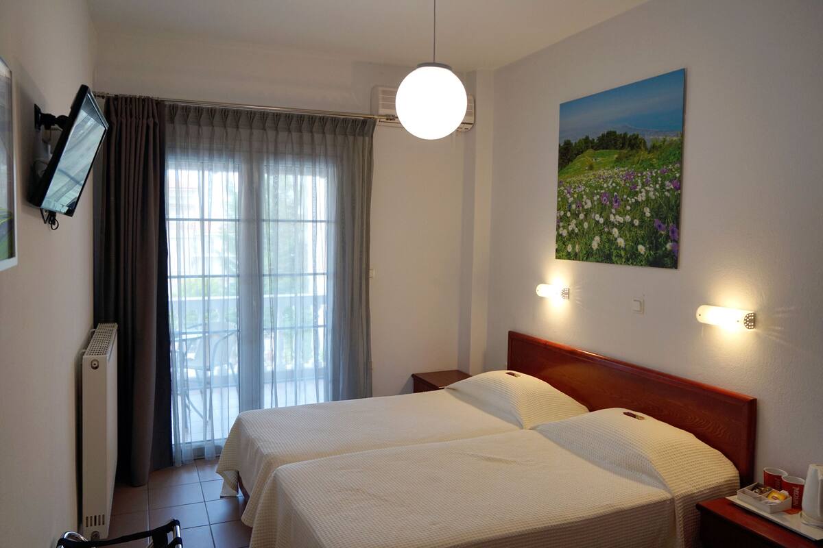 Hotelkamer met twee bedden, een groot raam met gordijnen, een schilderij aan de muur en een nachtkastje met een kopje en waterkoker.